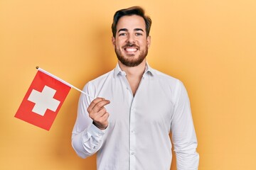 Wall Mural - Handsome caucasian man with beard holding switzerland flag looking positive and happy standing and smiling with a confident smile showing teeth