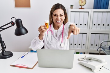 Canvas Print - Young hispanic woman wearing doctor uniform holding pills at the clinic pointing to you and the camera with fingers, smiling positive and cheerful