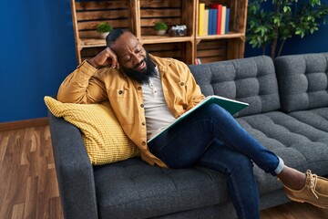 Sticker - Young african american man reading book sitting on sofa at home