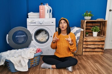 Wall Mural - Young hispanic woman doing laundry amazed and surprised looking up and pointing with fingers and raised arms.