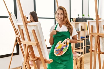 Wall Mural - Young hispanic middle age woman at art classroom pointing finger to one self smiling happy and proud