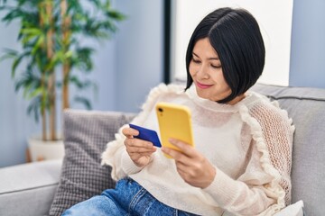 Wall Mural - Young chinese woman using smartphone and credit card sitting on sofa at home