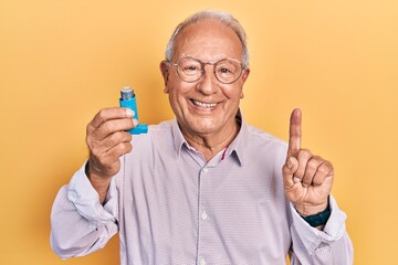 Wall Mural - Senior man with grey hair holding medical asthma inhaler surprised with an idea or question pointing finger with happy face, number one