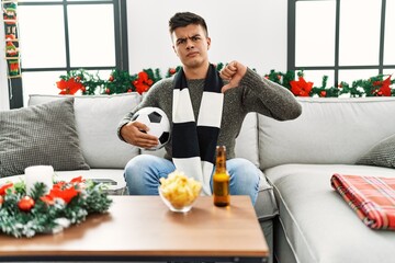 Poster - Young hispanic man football hooligan holding ball and beer with angry face, negative sign showing dislike with thumbs down, rejection concept