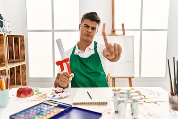 Sticker - Young hispanic man at art studio holding degree pointing with finger up and angry expression, showing no gesture