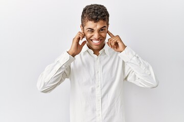 Sticker - Young handsome hispanic man standing over isolated background covering ears with fingers with annoyed expression for the noise of loud music. deaf concept.