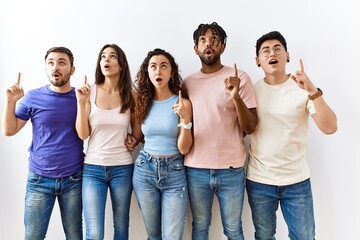 Canvas Print - Group of young people standing together over isolated background amazed and surprised looking up and pointing with fingers and raised arms.