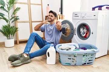 Wall Mural - Young hispanic man putting dirty laundry into washing machine pointing to you and the camera with fingers, smiling positive and cheerful