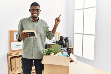 Canvas Print - Young african american businessman unboxing box at the office with a big smile on face, pointing with hand finger to the side looking at the camera.