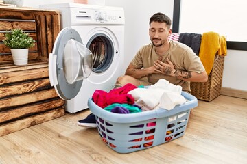 Canvas Print - Young handsome man putting dirty laundry into washing machine smiling with hands on chest with closed eyes and grateful gesture on face. health concept.