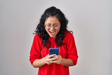 Canvas Print - Young asian woman using smartphone typing message angry and mad screaming frustrated and furious, shouting with anger. rage and aggressive concept.