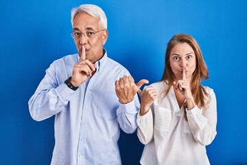 Canvas Print - Middle age hispanic couple standing over blue background asking to be quiet with finger on lips pointing with hand to the side. silence and secret concept.