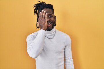 Wall Mural - African man with dreadlocks wearing turtleneck sweater over yellow background covering one eye with hand, confident smile on face and surprise emotion.