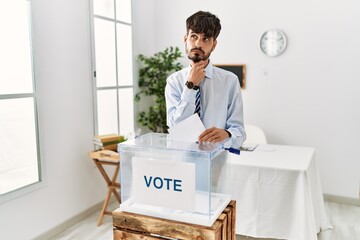 Wall Mural - Hispanic man with beard voting putting envelop in ballot box with hand on chin thinking about question, pensive expression. smiling with thoughtful face. doubt concept.