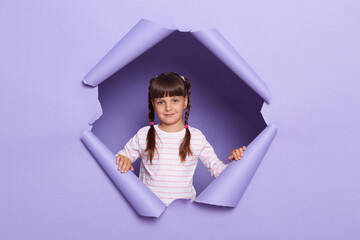 Wall Mural - Portrait of positive happy little girl wearing striped shirt posing in torn purple paper hole, expressing optimism, looking at camera with calm expression.