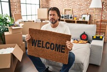 Handsome Middle Age Man Holding Welcome Doormat At New Home Skeptic And Nervous, Frowning Upset Because Of Problem. Negative Person.