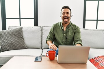 Poster - Young hispanic man with beard wearing call center agent headset working from home sticking tongue out happy with funny expression. emotion concept.