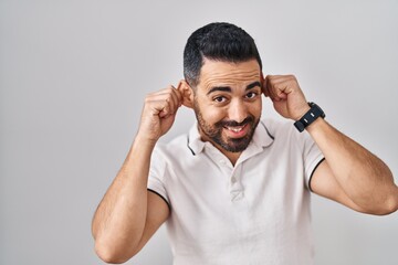 Poster - Young hispanic man with beard wearing casual clothes over white background smiling pulling ears with fingers, funny gesture. audition problem