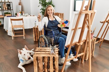 Poster - Caucasian young blonde woman sitting on wheelchair at art studio smiling happy pointing with hand and finger to the side