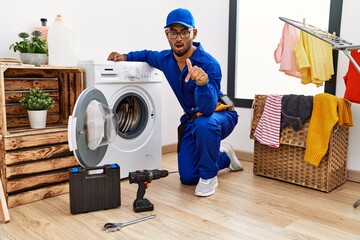 Poster - Young indian technician working on washing machine pointing down with fingers showing advertisement, surprised face and open mouth