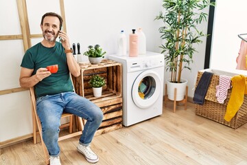 Wall Mural - Middle age hispanic man doing laundry talking on the smartphone and drinking coffee at home.