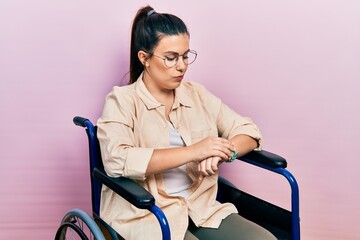 Poster - Young hispanic woman sitting on wheelchair checking the time on wrist watch, relaxed and confident