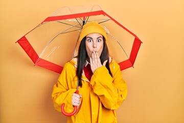 Poster - Young hispanic woman wearing yellow raincoat holding umbrella covering mouth with hand, shocked and afraid for mistake. surprised expression