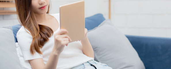 Wall Mural - Young attractive asian woman resting using browsing tablet computer on sofa at home, happy girl sitting on couch relax reading digital gadget at house, communication and lifestyle concept.