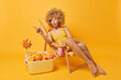 canvas print picture - Shocked curly haired young woman dressed in swimsuit points left with index finger poses on comfortable deck chair near portable freezer with citrus fruits isolated over vivid yellow background