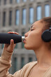 Vertical shot of thirsty young woman leads sporty lifestyle drinks fresh water from bottle dressed in sweatshirt uses stereo headphones feels tired stands outdoors against blurred background