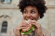 © wayhome.studio  - Pretty curly haired woman bites delicious sandwich poses outdoors at street looks away dressed casually has quick snack while walking outside being hungry. People lifestyle and fast food concept