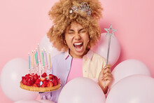 Positive Woman With Curly Hair Wears Crown On Head Holds Magic Wand Exclaims Loudly Holds Delicious Strawberry Cake Poses Around Inflated Balloons Isolated Over Pink Background. Festivity Concept