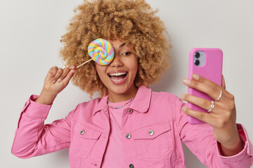 Wall Mural - Positive woman covers eye with sweet lollipop laughs gladfully takes selfie via smartphone dressed in fashionable pink jacket isolated over grey background. Smiling girl holds delicious caramel candy