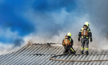 Firefighters Extinguish Flame On Roof Of Building. Roofing Houses Covered Fire. Rescuers Pour Fire With Water From Hose. Emergency Concept. Firefighters In Uniform. Fire On Roof Of House.