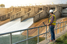 A Dam Engineering Doing His Checking Routine. He Is Wearing A White Hard Hat And Yellow Transparent Vest. He Is Standing By The Rail By The Dam.