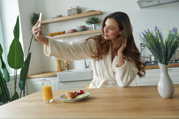 Sticker - Attractive young woman in bathrobe making selfie while having breakfast at the modern kitchen