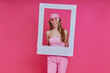 © gstockstudio - Happy young woman looking through a picture frame while standing against pink background