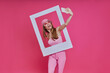 canvas print picture - Playful young woman making selfie and looking through a picture frame while standing against pink background
