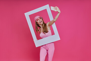playful young woman making selfie and looking through a picture frame while standing against pink ba