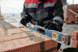 © pressmaster - Gloved hands of male constructor or foreman in workwear holding level on surface of brick wall at construction site while checking flatness