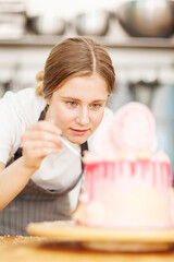 Young female pastry chef decorating defocused handmade cake placed on rotating stand in bakery shop kitchen