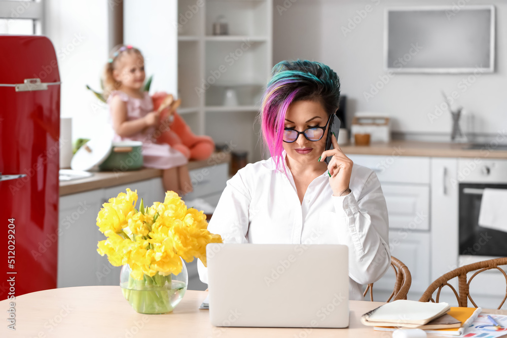 Mother working with laptop and mobile phone at table while her little daughter playing in kitchen