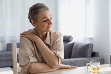 Mature Woman Sitting At Table At Home With Closed Eyes Hugging Herself