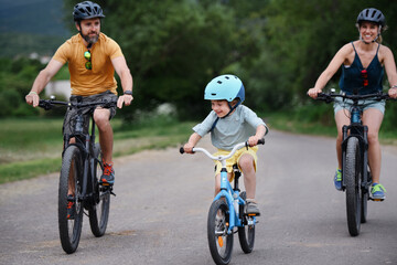 Young family with little child riding bicycles on road in village in summer.