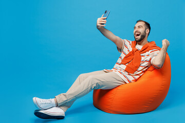 Wall Mural - Full body young overjoyed happy man in orange striped t-shirt sit in bag chair doing selfie shot on mobile cell phone post photo on social network do winner gesture isolated on plain blue background.