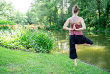 Young Caucasian Woman Standing In Yoga Pose On One Leg With Hands On Back As In Prayer Meditating, Working Out Or Pilates On Green Grass Loan Near Calm River Or Lake In Summer Park. Back View, No Face
