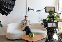 Behind The Scenes On Video Shoot Of Man Being Interviewed On Camera. Caucasian Business Man Sitting On Couch With Microphone, Lighting And Camera In Frame.