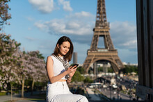 Young Woman In Stylish Outfit Using Smartphone While Sitting Near Eiffel Tower In Paris.