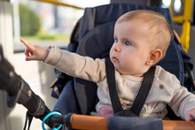 A Cute Little Baby Of 10 Months Is Sitting In A Stroller. A Child Is Fastened With Seat Belts In Stroller Rides A Passenger Bus With Her Mother. Concept Of Child Safety.