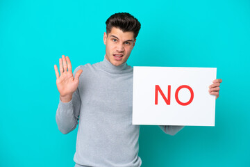 Young handsome caucasian man isolated on blue bakcground holding a placard with text NO and doing stop sign